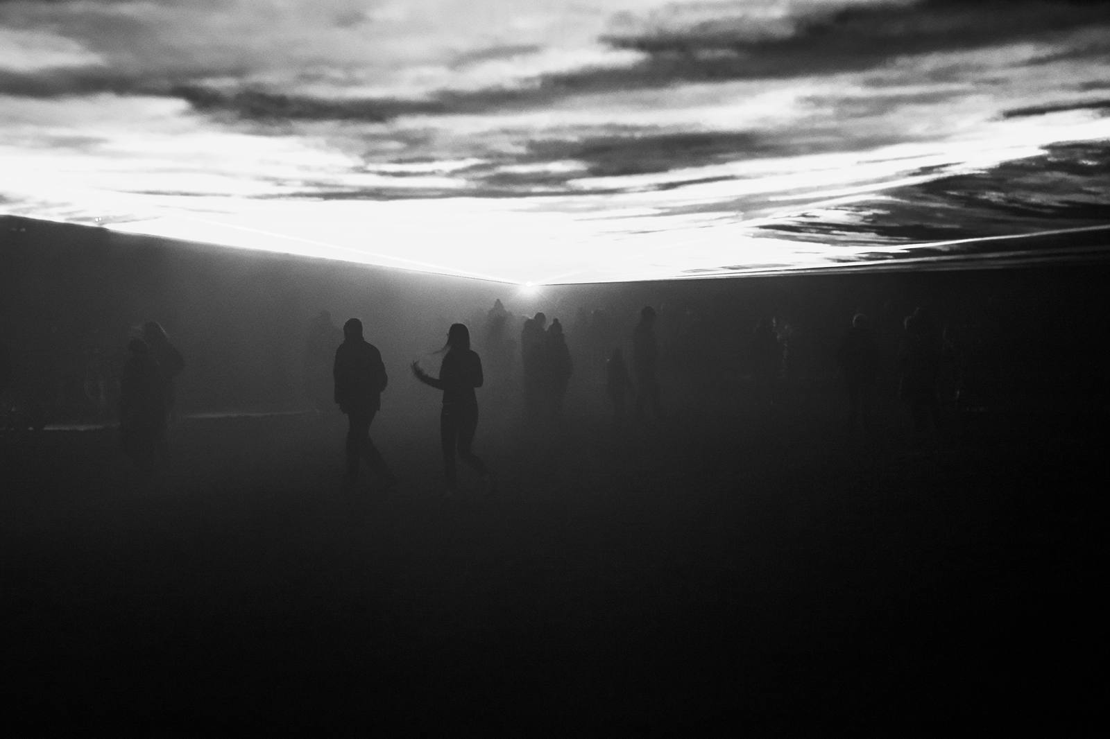 People walk through fog at dusk beneath a flat sheet of light.